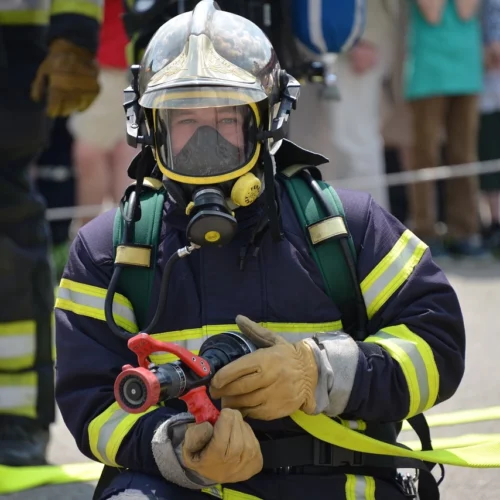 Feuerwehrmänner in voller Montur, vordere Person hält Feuerwehrspritze